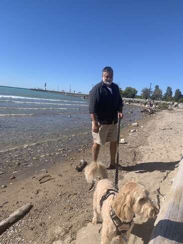 Ein Mann mit Bart steht an einem Sandstrand und hält einen flauschigen hellbraunen Hund an der Leine. See, blauer Himmel und Pier im Hintergrund.
