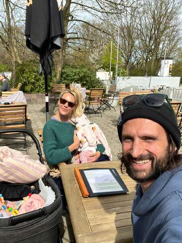 Un homme sourit pour un selfie à la terrasse d'un café. Une femme portant des lunettes de soleil tient un bébé dans ses bras. On aperçoit une poussette et une tablette sur la table.