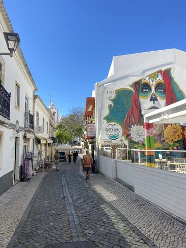 Une ruelle pavée bordée de bâtiments blancs, des œuvres de street art colorées représentant un visage du Jour des Morts, des terrasses de café et un ciel d'un bleu éclatant.