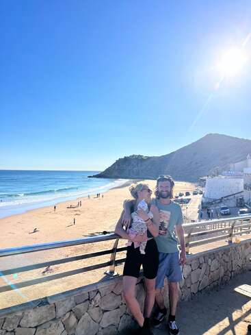 Un couple souriant se tient avec son bébé sur un belvédère en bord de mer, baigné de soleil. L'océan, le sable doré, les montagnes et le ciel d'un bleu éclatant composent une scène joyeuse et pittoresque.