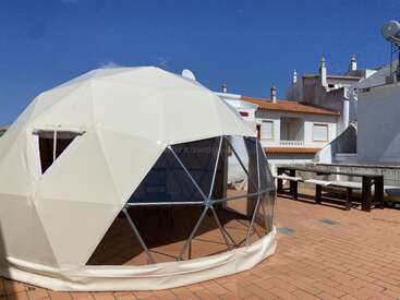 Une tente en forme de dôme géodésique blanche est installée sur une terrasse carrelée. Elle est entourée de bâtiments blancs de style méditerranéen aux toits orange, sous un ciel bleu clair et lumineux.