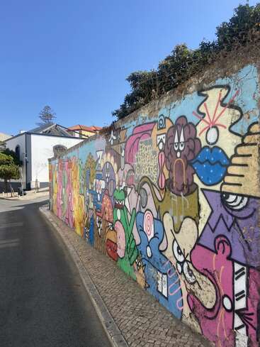 Une fresque murale de graffiti, vive et colorée, recouvre un long mur dans une rue ensoleillée. Des personnages de dessins animés espiègles et des formes abstraites créent une ambiance animée et artistique près du trottoir.