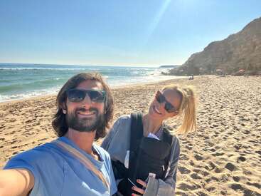 Un couple souriant, tous deux portant des lunettes de soleil, prend un selfie sur une plage ensoleillée. La femme porte un bébé dans un porte-bébé ventral. On aperçoit l'océan, le sable et les falaises.