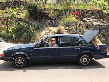 Une femme conduit une Volvo bleu foncé avec un gros tas de foin à l'arrière, garée sur le bord d'une route devant une clôture à mailles losangées.