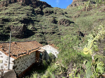 L'image représente une petite maison rustique au toit de tuiles située dans une zone montagneuse, entourée de verdure et de cactus.