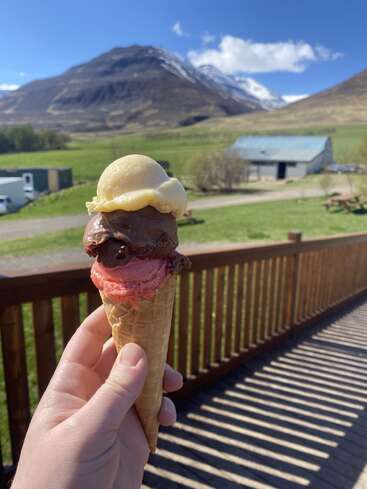 A hand holds a triple-scoop ice cream cone outdoors, with beautiful mountains, clear blue sky, and a rustic barn in the scenic background. Sunny, peaceful day.