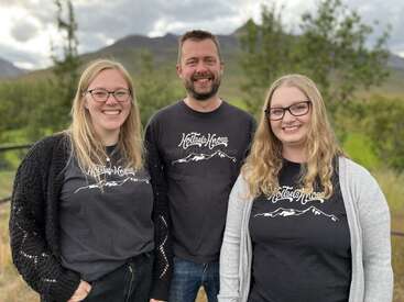 Three smiling people stand outdoors, all wearing glasses and matching “Holtsel Knoss” shirts. The background features green trees, mountains, and a cloudy sky. They look happy.