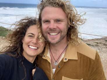 A happy couple smiles broadly while taking a selfie at a windy, scenic beach. The ocean waves crash behind them, and their curly hair blows freely.