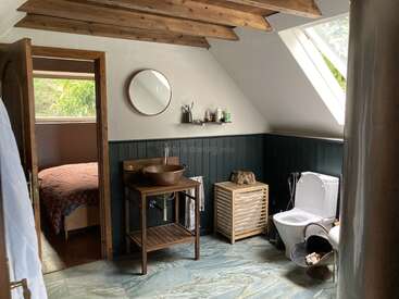 Cette salle de bains confortable est dotée de poutres rustiques en bois, d'une lucarne, d'un lavabo vasque, de toilettes modernes et d'un panier de rangement. Une chambre attenante avec une fenêtre est également visible.