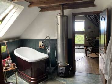 Cette salle de bain confortable comprend une baignoire vintage, des murs lambrissés, un grand chauffe-eau métallique, un plafond incliné et une pièce attenante dotée de grandes fenêtres laissant entrer la lumière du soleil.