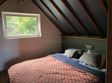 Cette chambre mansardée confortable dispose d'un plafond incliné avec poutres apparentes, d'une grande fenêtre avec vue sur la verdure, d'un lit double et d'une literie sombre et moelleuse.