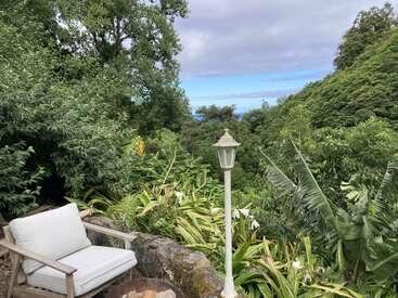 Une chaise d'extérieur confortable est placée près d'un foyer en pierre, au milieu d'une végétation luxuriante, avec un lampadaire blanc et un ciel bleu lointain à l'horizon.