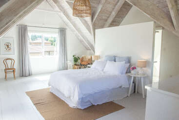 La imagen representa un dormitorio sereno con una cama grande, una cómoda y una ventana, con el suelo de madera clara y el techo blanco, que transmite una sensación de calma y tranquilidad.