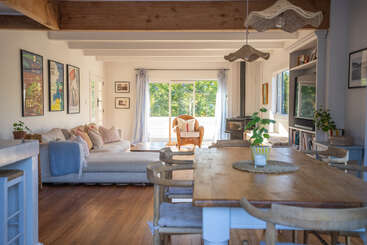 Este acogedor salón-comedor cuenta con una mesa de madera, un cómodo sofá, paredes llenas de arte, luz natural, plantas, cortinas suaves y un sillón de mimbre junto a grandes ventanales.