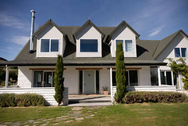 Esta imagen muestra una casa moderna de dos plantas con tejado gris oscuro y exterior blanco, grandes ventanales, porche delantero y jardín paisajista bajo un cielo azul.