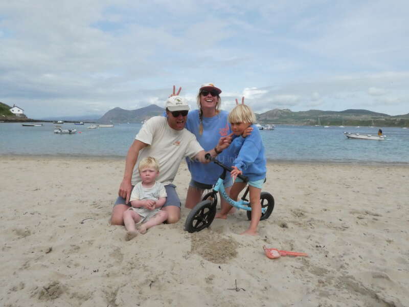 La imagen muestra a una familia de cuatro miembros posando en una playa de arena, con una serena masa de agua y barcos al fondo, y un cielo nublado.