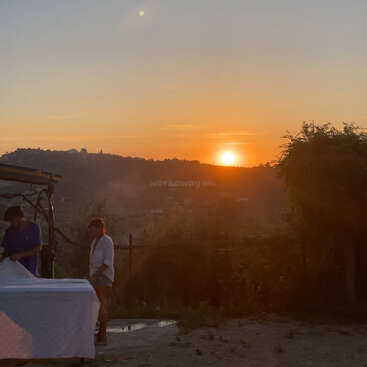 Dos personas están al aire libre cerca de una mesa al atardecer, con el sol dorado poniéndose sobre colinas y árboles distantes, proyectando un cálido resplandor sobre el paisaje.