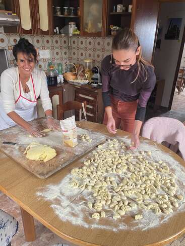 Two women are making homemade pasta in a cozy kitchen. Dough, flour, and many pieces of fresh pasta cover the wooden table as they smile and work.
