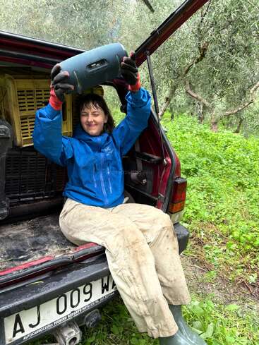 A person in work clothes and gloves sits on the back of a red vehicle, holding a speaker above their head, surrounded by lush green vegetation.