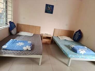 Esta imagen muestra una habitación sencilla con dos camas individuales, cada una con una almohada y una manta con motivos florales, una mesita de noche, una ventana y un cuadro en la pared.