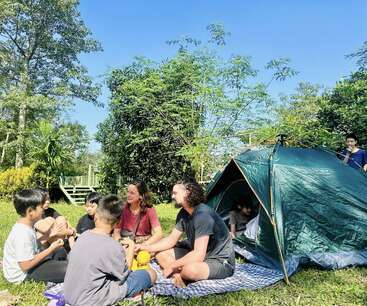 Un grupo de personas, entre las que hay adultos y niños, está sentado sobre una manta de picnic junto a una tienda de campaña verde al aire libre, riendo y charlando bajo un cielo azul brillante.