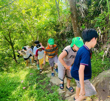 Un grupo de niños sube por un sendero estrecho y rocoso, rodeado de frondosos árboles y plantas, disfrutando juntos de un día lleno de aventuras al aire libre en plena naturaleza.
