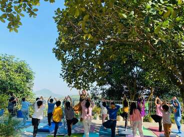 Eine Gruppe von Kindern macht unter einem großen Baum im Freien Yoga und folgt dabei einem Lehrer. Bunte Matten und üppiges Grün sorgen für eine friedliche, lebendige und gesunde Atmosphäre.