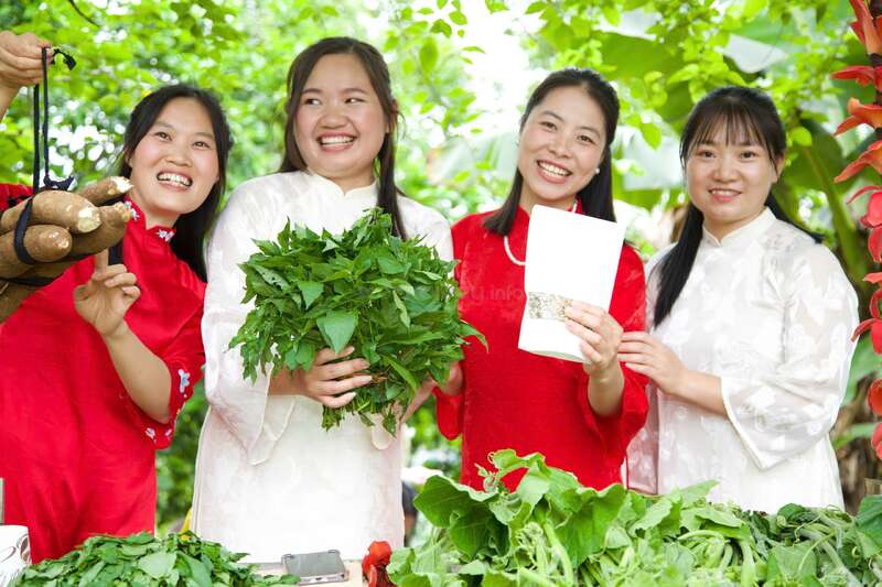 Tres mujeres, vestidas con el tradicional áo dài vietnamita, sonríen felices al aire libre, rodeadas de una exuberante vegetación. Una sostiene un ramo de hojas, otra lleva un sobre blanco. Ambiente festivo.