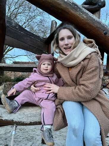 A woman and a baby, both dressed warmly, sit on a wooden structure at a playground, smiling. Trees and buildings are visible in the background.