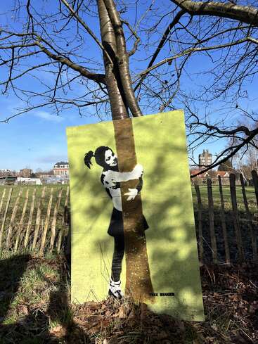 A painted board leans against a tree, depicting a girl hugging the trunk. The background is green, with clear skies and wooden fencing nearby, labeled "Tree Hugger."