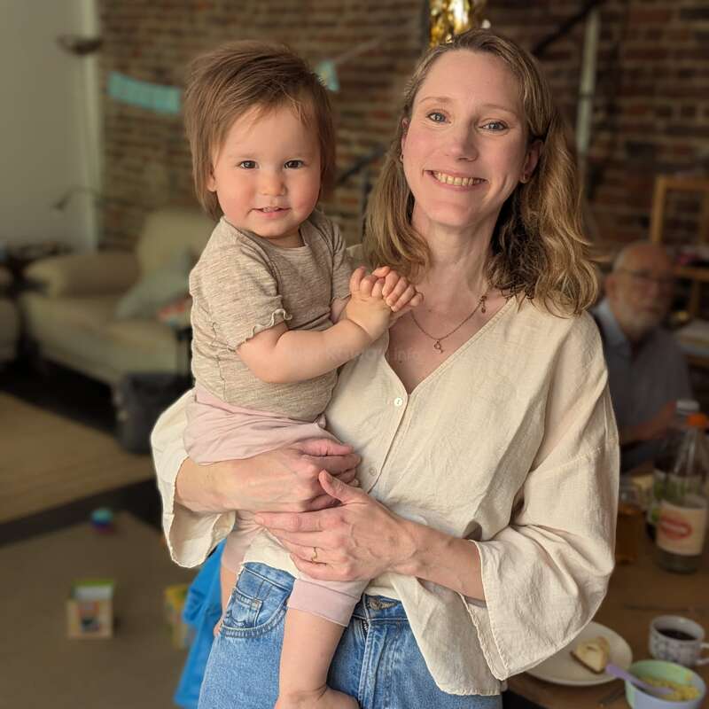 A smiling woman holds a cheerful toddler in a cozy, warmly lit room. There’s a man in the background, and homey details add comfort and warmth.