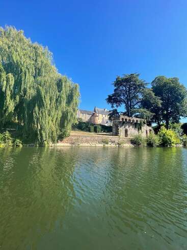 A serene river reflects lush green trees, an ancient stone building, and distant historic structures under a clear, vibrant blue sky on a sunny day.
