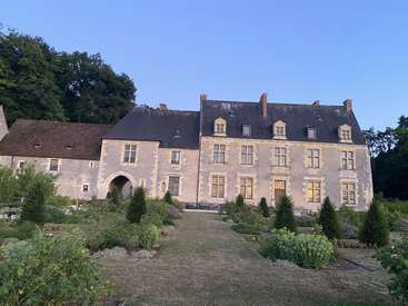 A historic stone manor stands surrounded by lush gardens, neatly trimmed bushes, and vibrant greenery. The evening light casts a peaceful glow over the scene.
