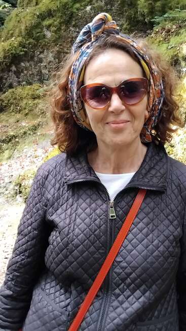 A woman with curly hair, wearing sunglasses, a colorful headscarf, and a quilted jacket, stands outdoors in front of a mossy, rocky background, smiling gently.
