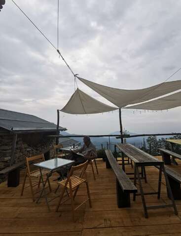 A person sits alone reading on a rustic wooden deck with tables, chairs, and sunshades overhead. Mountains and cloudy sky create a peaceful, serene atmosphere.