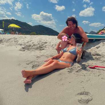 A family enjoys a sunny beach day. A woman in a bikini relaxes with a drink, a baby with a flower sticker, and a man smiling.