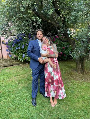 A couple stands outdoors, dressed elegantly, holding a baby with a pink flower on its face. They are surrounded by lush greenery and blooming hydrangeas.