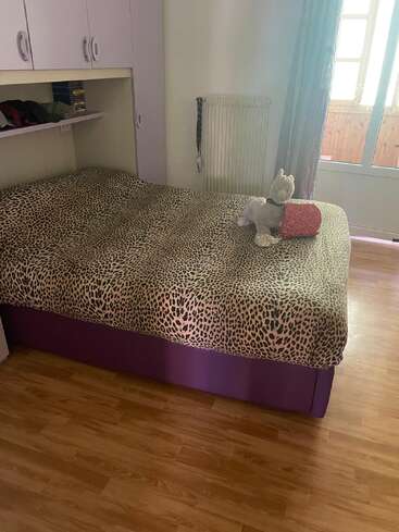 A cozy bedroom with a cheetah-print bedspread, plush toy, purple bed base, wooden floor, white cabinets, shelf with books, window, and radiator visible.