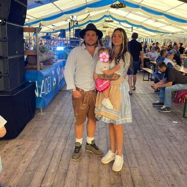A family dressed in traditional Bavarian attire stands inside a festive beer tent, surrounded by people seated at tables, enjoying a lively event with decorations overhead.
