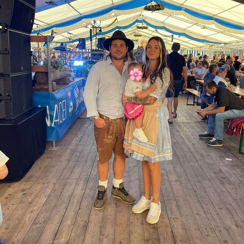A family dressed in traditional Bavarian attire stands inside a festive beer tent, surrounded by people seated at tables, enjoying a lively event with decorations overhead.