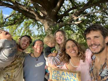 Un alegre grupo de siete personas, incluidos niños, permanecen juntos al aire libre bajo un gran árbol, sonriendo y sosteniendo un cartel de madera en el que se lee "CRANN BETHADH".