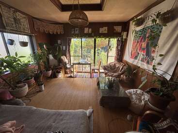 This cozy living room features abundant potted plants, woven decor, a coffee table, art tapestry, large windows, natural light, and a small table with chairs.