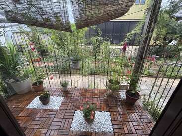This image shows a cozy patio with wooden tiles, potted plants, and white pebbles, overlooking a lush, green garden with various plants and a bamboo shade.
