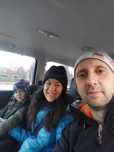 Das Bild zeigt eine dreiköpfige Familie auf dem Rücksitz eines Autos, in Winterkleidung, mit einem Mann, einer Frau und einem Kind, die alle in die Kamera lächeln.