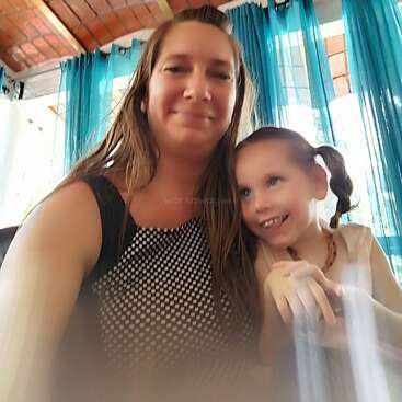 A imagem mostra uma mulher e uma menina posando para uma selfie em uma sala com teto de azulejos, cortinas azuis e uma janela atrás delas.