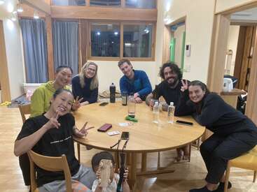 Seis adultos e uma criança sentam-se ao redor de uma mesa de madeira, sorrindo e fazendo sinais de paz. Garrafas de água, cartões e telefones estão sobre a mesa em uma sala aconchegante.