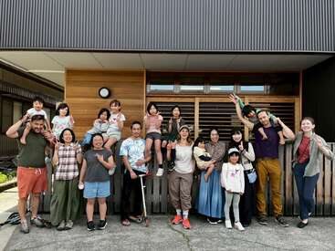 Um grupo diversificado de adultos e crianças posa alegremente do lado de fora de um edifício moderno, sorrindo e fazendo sinais de paz, demonstrando amizade e união em um dia ensolarado.