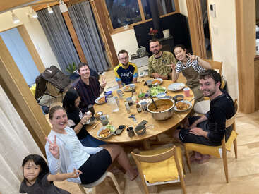 Um grupo de amigos e familiares desfruta de uma refeição juntos em torno de uma mesa de madeira. Eles estão sorrindo, fazendo sinais de paz e compartilhando alimentos em uma casa aconchegante.