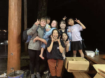 Um grupo de sete amigos, incluindo crianças e adultos, posam alegremente juntos à noite, piscando sinais de paz, cercados por postes de madeira, mesas de piquenique e suprimentos.