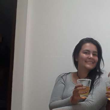 A imagem mostra uma mulher com longos cabelos escuros, vestindo uma camisa cinza-clara, segurando um copo com um líquido amarelo em frente a uma parede branca.